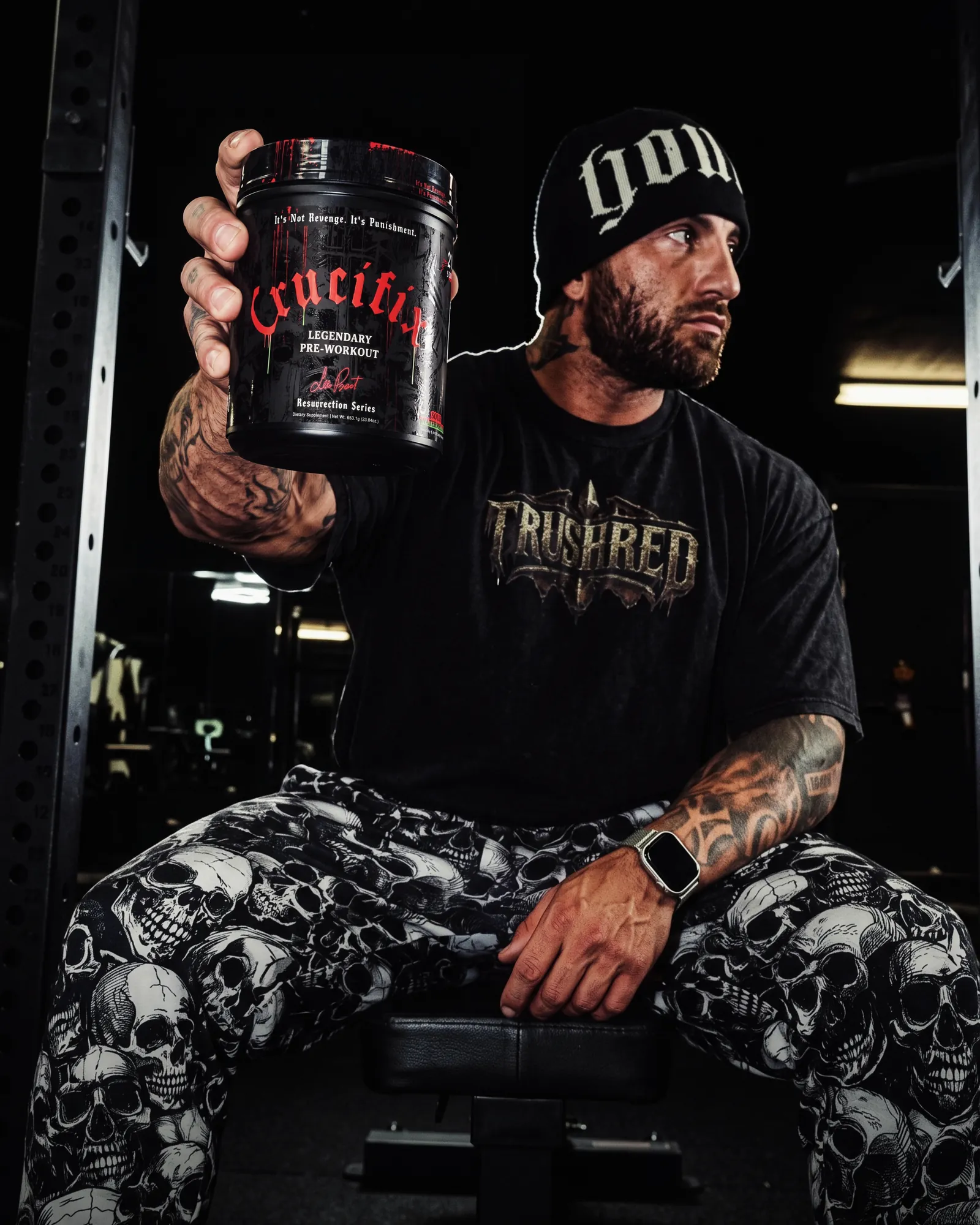 Tattooed athlete in skull-print pants raising a NutriFitt Crucifix Legendary Pre-Workout tub toward the camera in a dark gym.