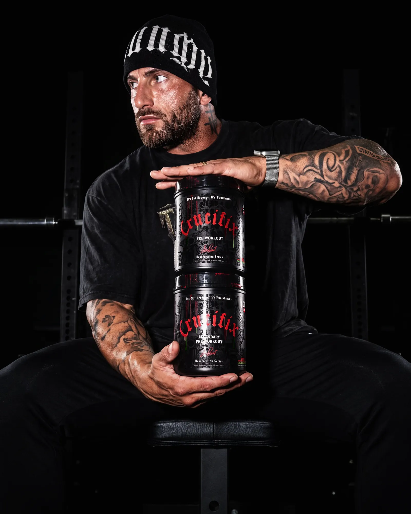 Tattooed athlete in a beanie seated on a gym bench holding two stacked NutriFitt Crucifix pre-workout tubs.
