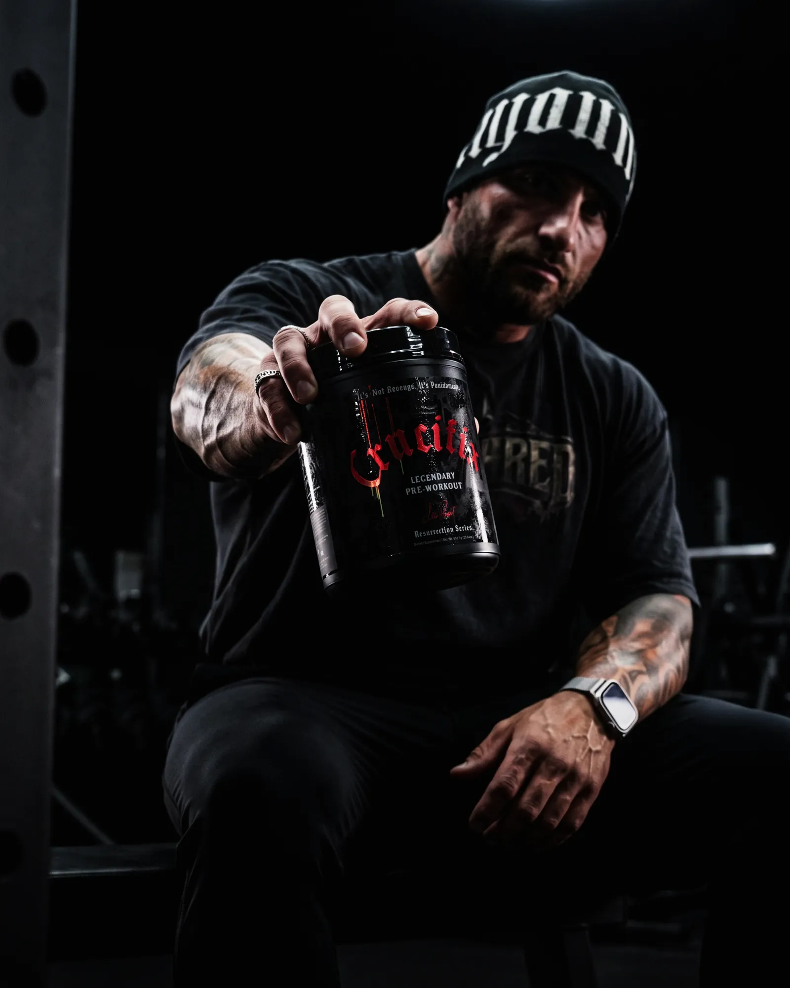 Seated athlete in a dark gym holding a NutriFitt Crucifix Legendary Pre-Workout tub out toward the camera.