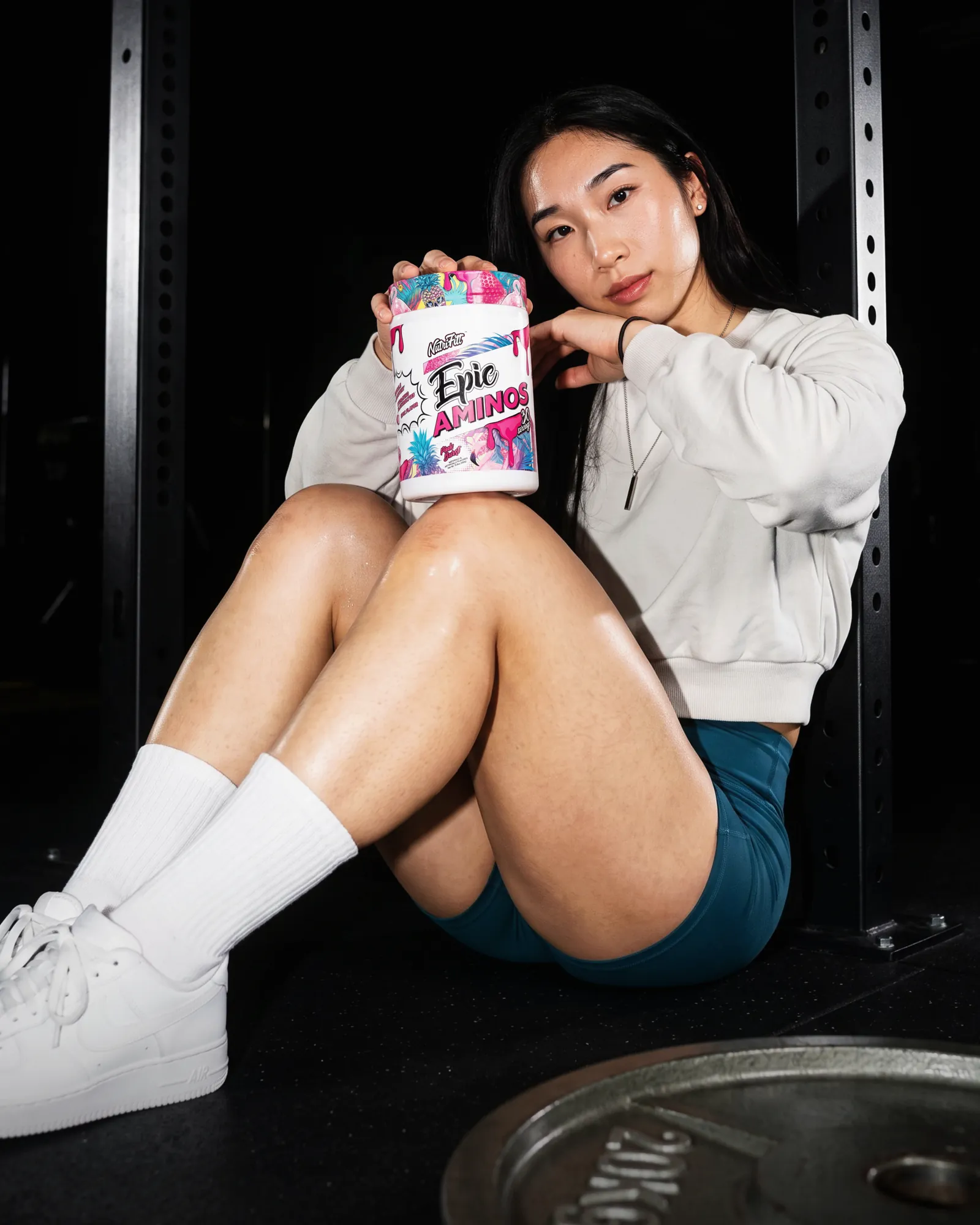 Athlete in a cream sweatshirt seated on a gym floor, resting a NutriFitt Epic Aminos tub on her knee.