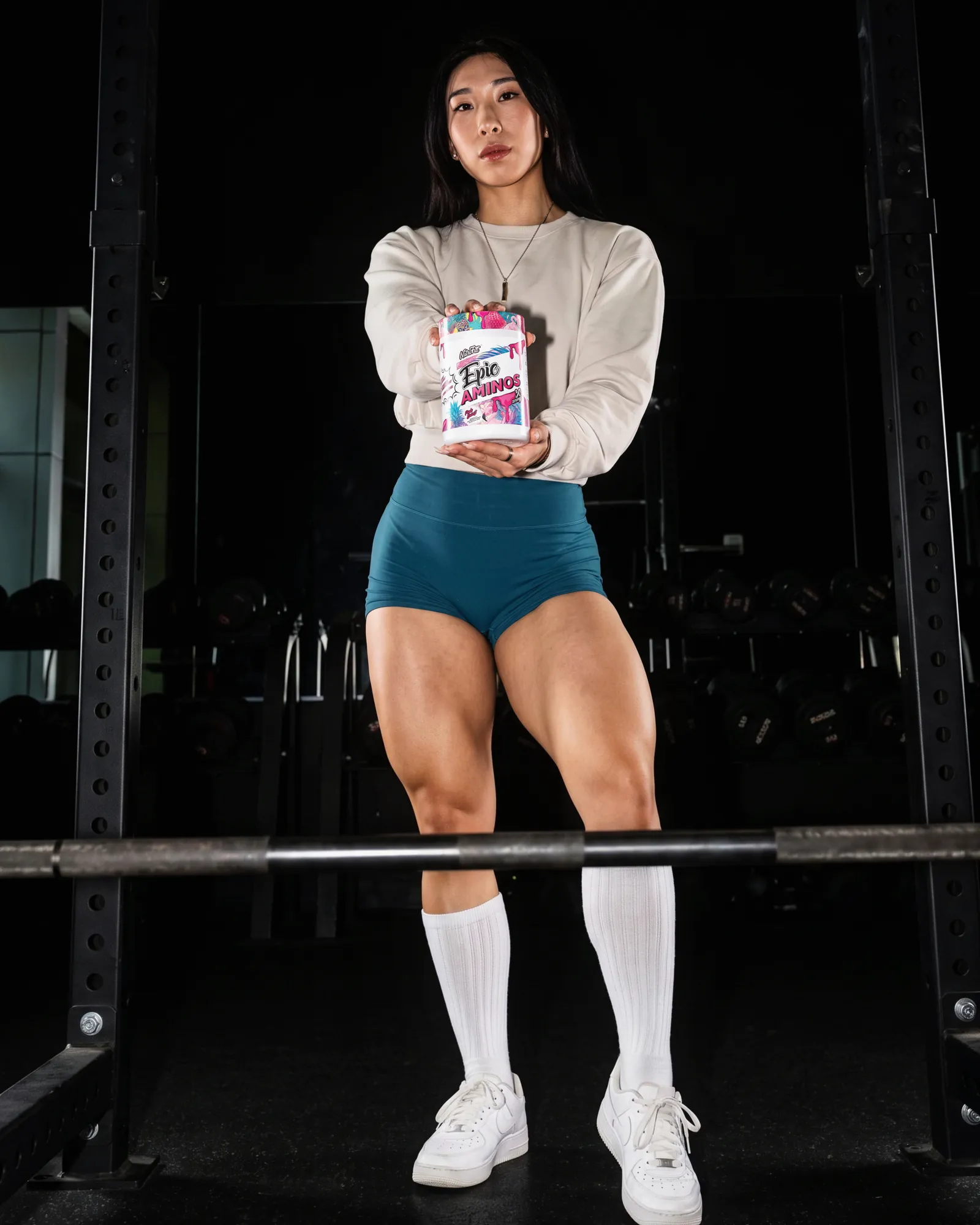 Athlete standing inside a squat rack holding a NutriFitt Epic Aminos tub out toward the camera.