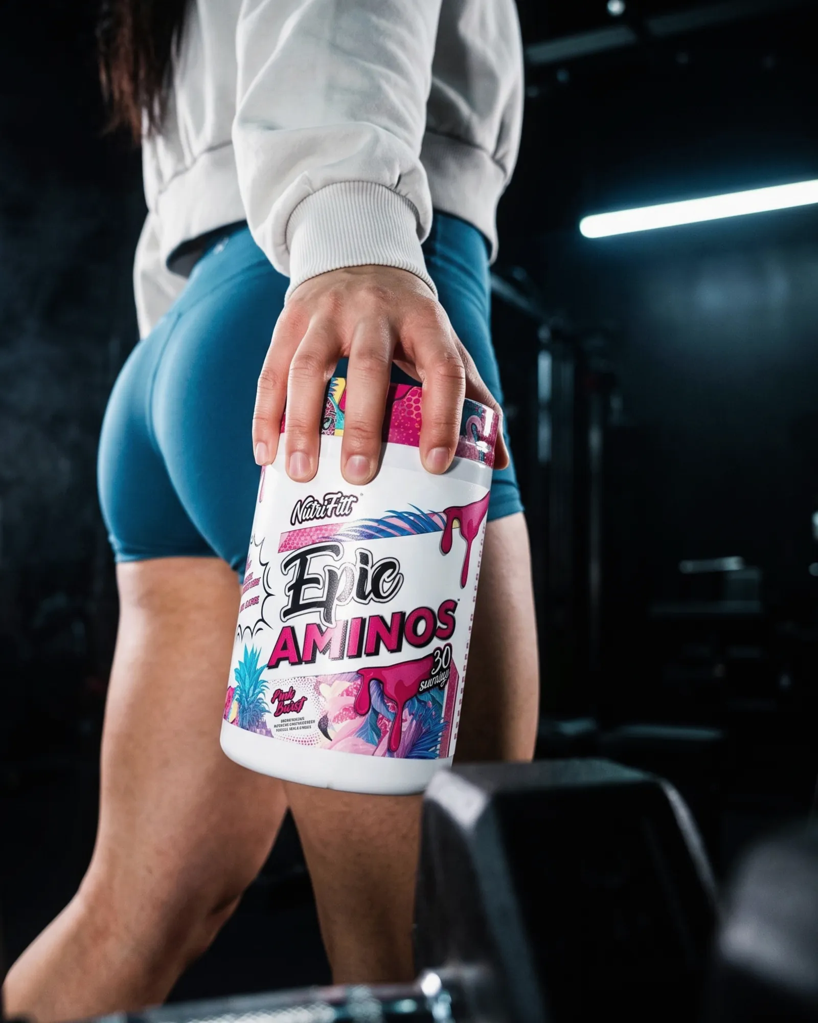 Low-angle view of an athlete holding a NutriFitt Epic Aminos tub against her leg in a dark gym.