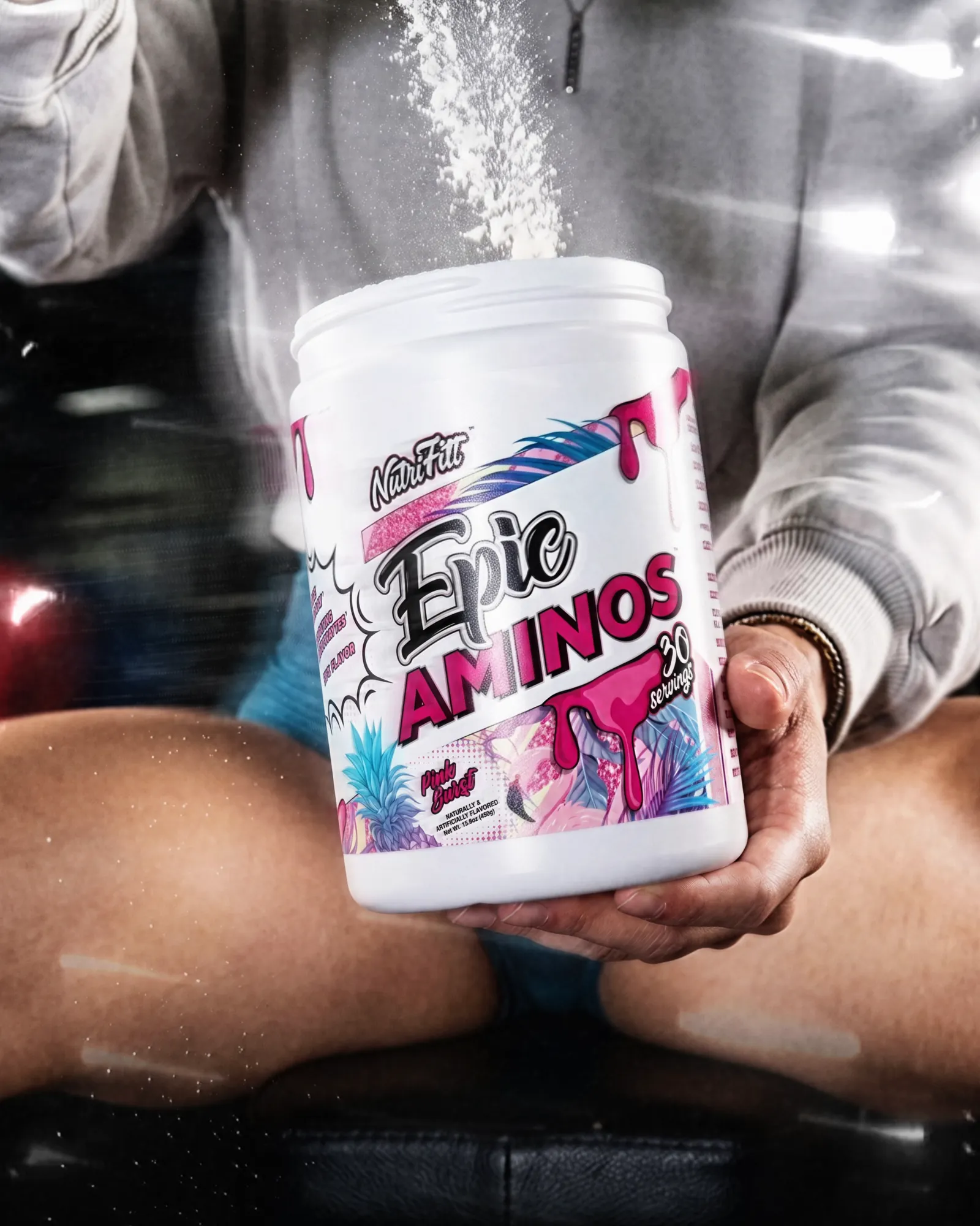 NutriFitt Epic Aminos tub held in both hands as powder falls, shot through glass with light reflections.