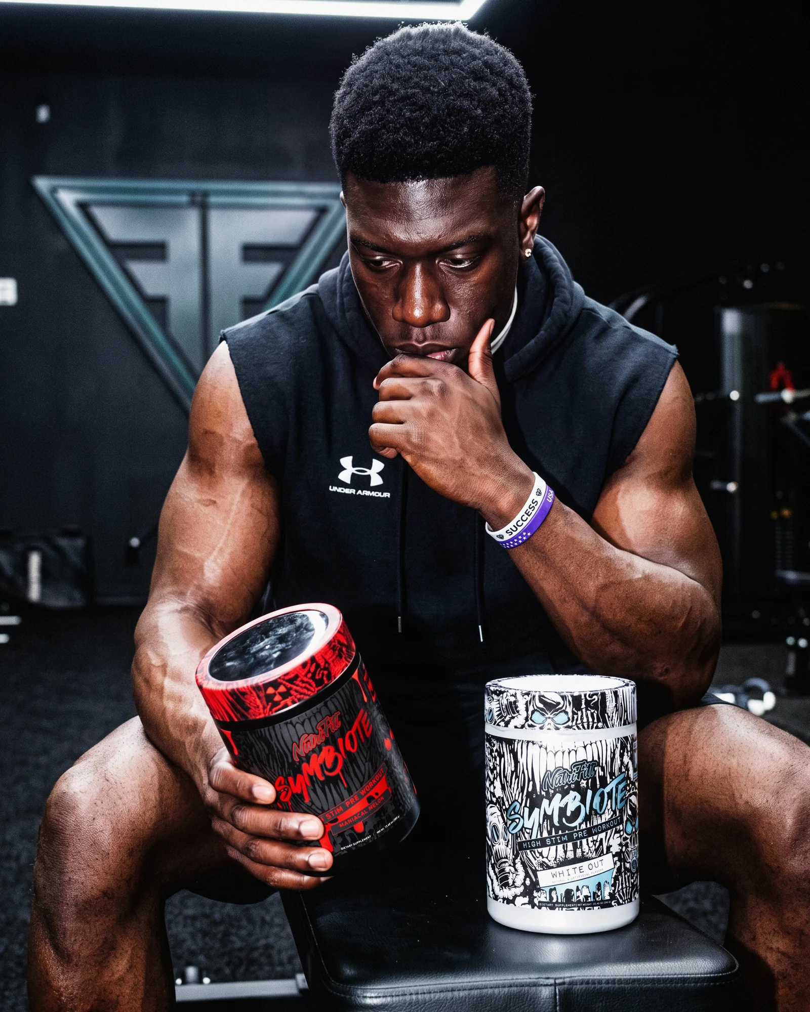 Seated athlete holding a red NutriFitt Symbiote tub beside a white Symbiote White Out tub in a dark gym.