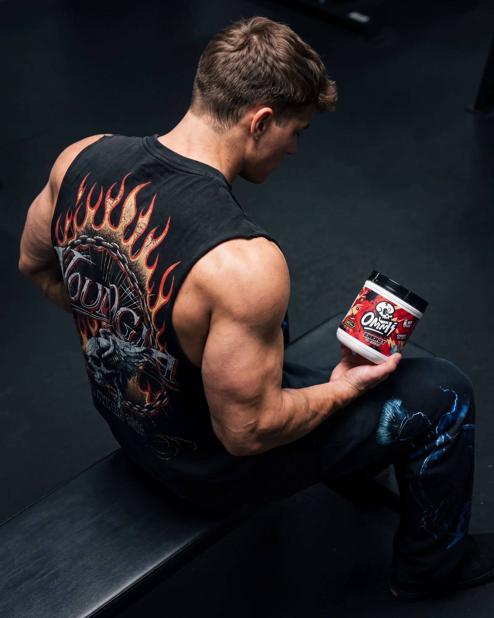 Muscular athlete seated on a gym bench holding Ommi Energy Cherry Pop Cream pre-workout.