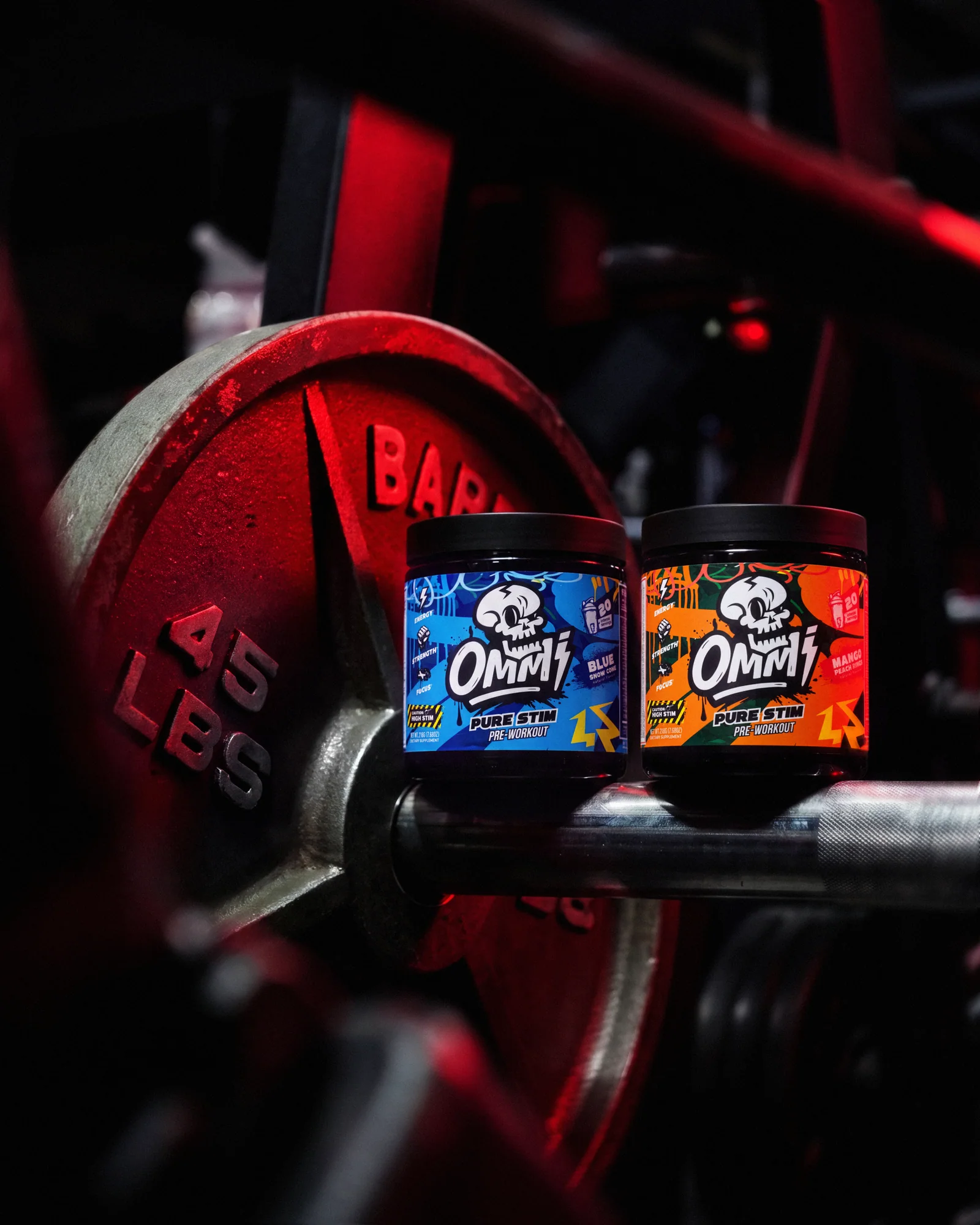 Ommi Pure Stim Blue Snow Cone and Mango Peach Rings pre-workout tubs beside gym weights under red light.