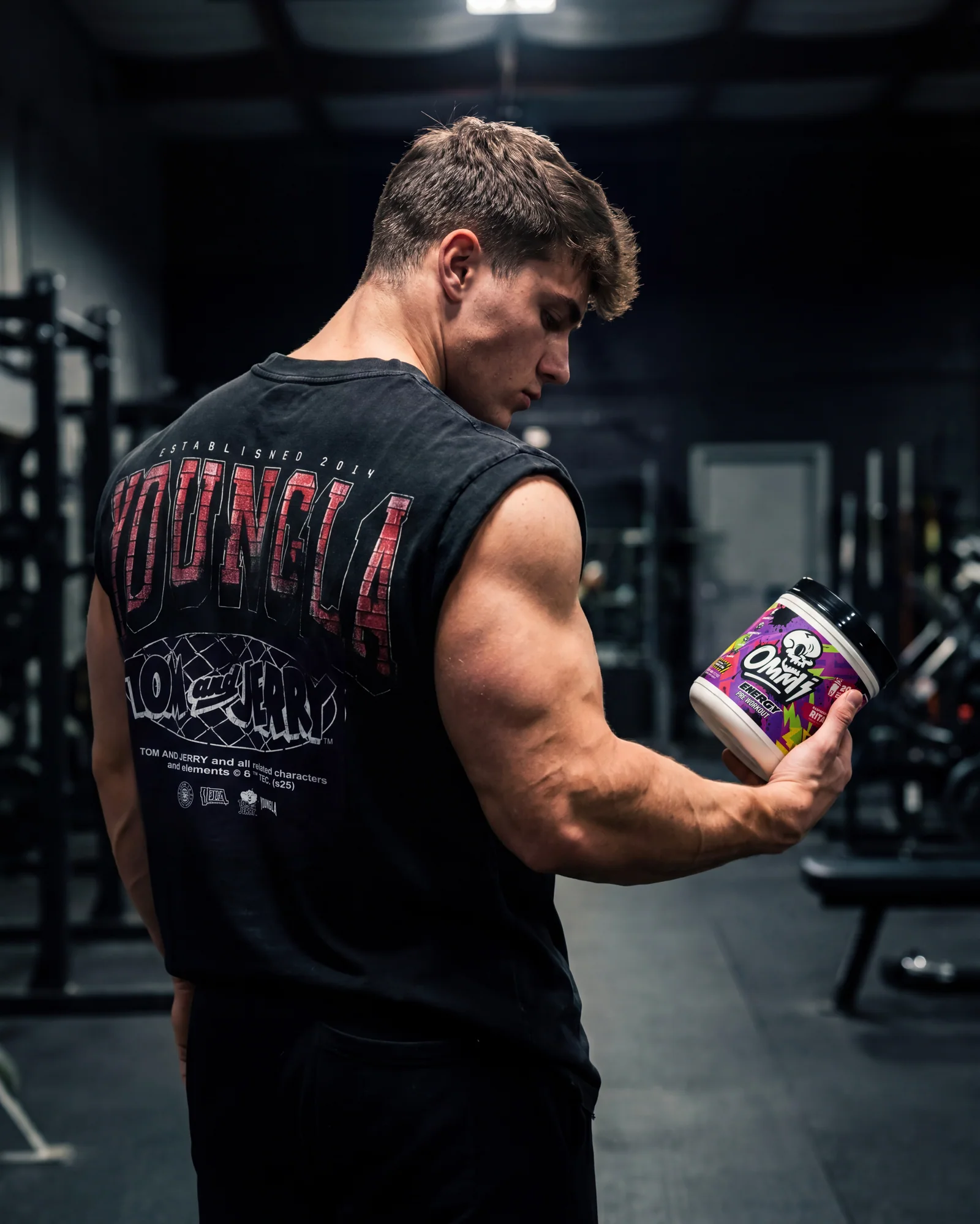 Muscular athlete in a dark gym holding an Ommi Energy Raspberry Rita pre-workout tub.