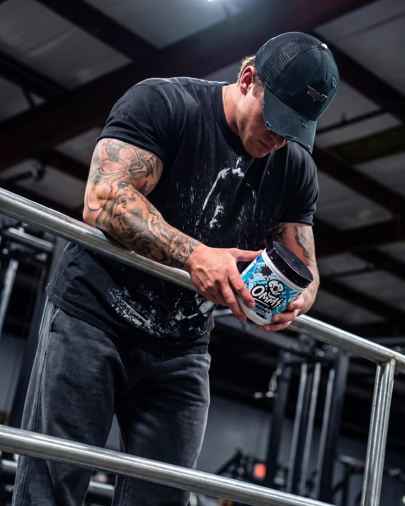 Tattooed athlete in a black cap holding a blue Ommi Energy pre-workout tub beside gym rails.