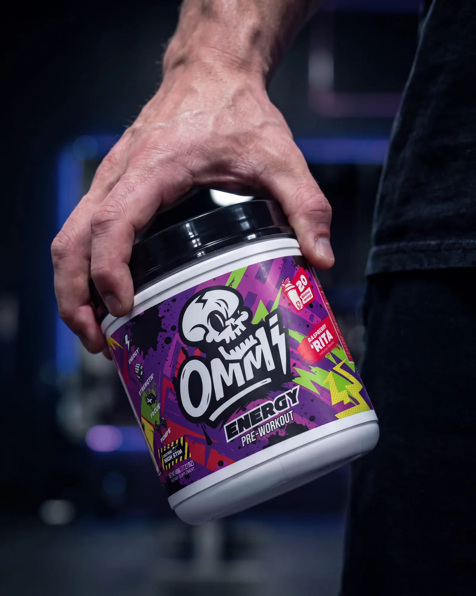 Close-up of a hand gripping an Ommi Energy Raspberry Rita pre-workout tub in a dark gym.