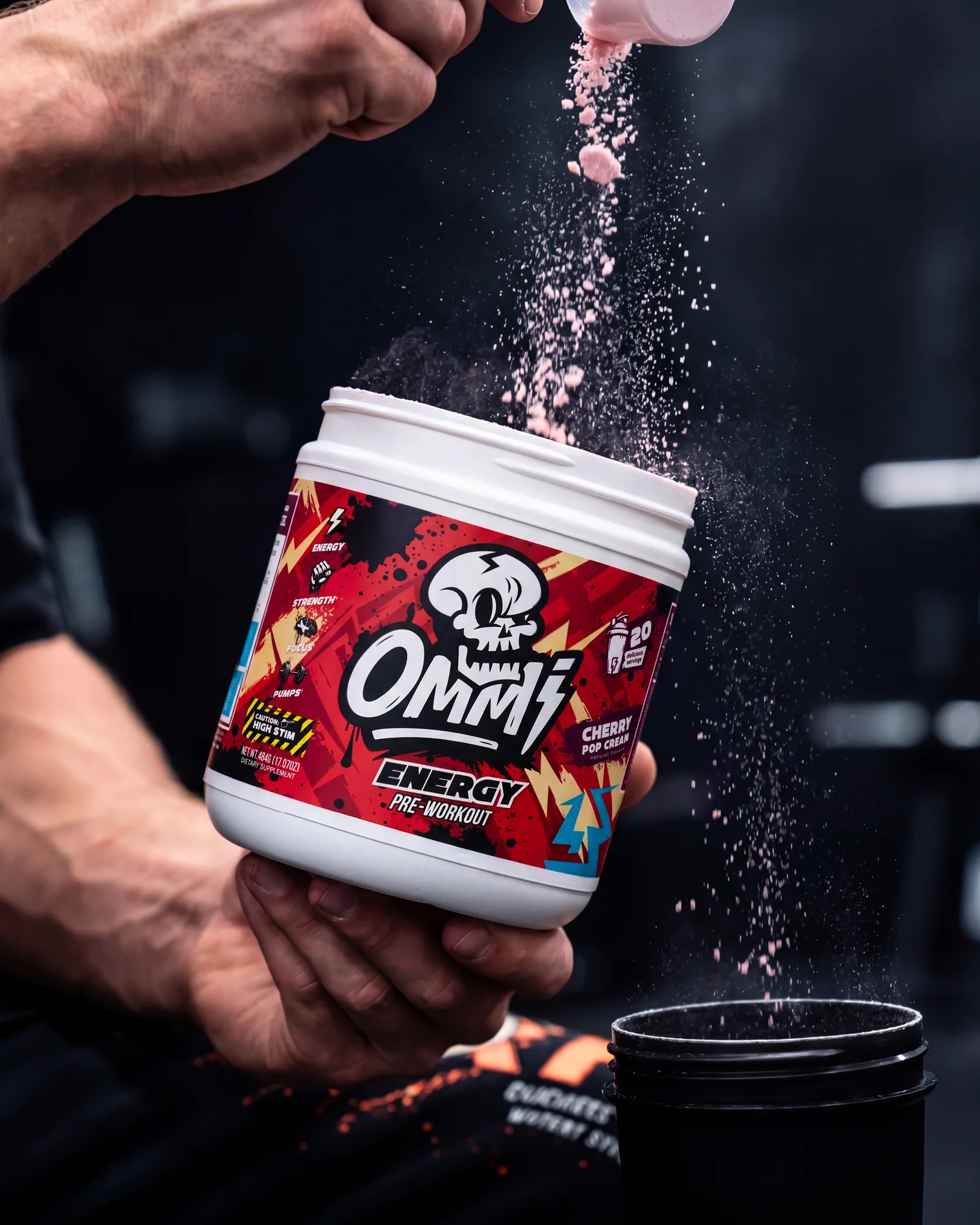 Close-up of pink pre-workout powder being poured beside an Ommi Energy Cherry Pop Cream tub and shaker.
