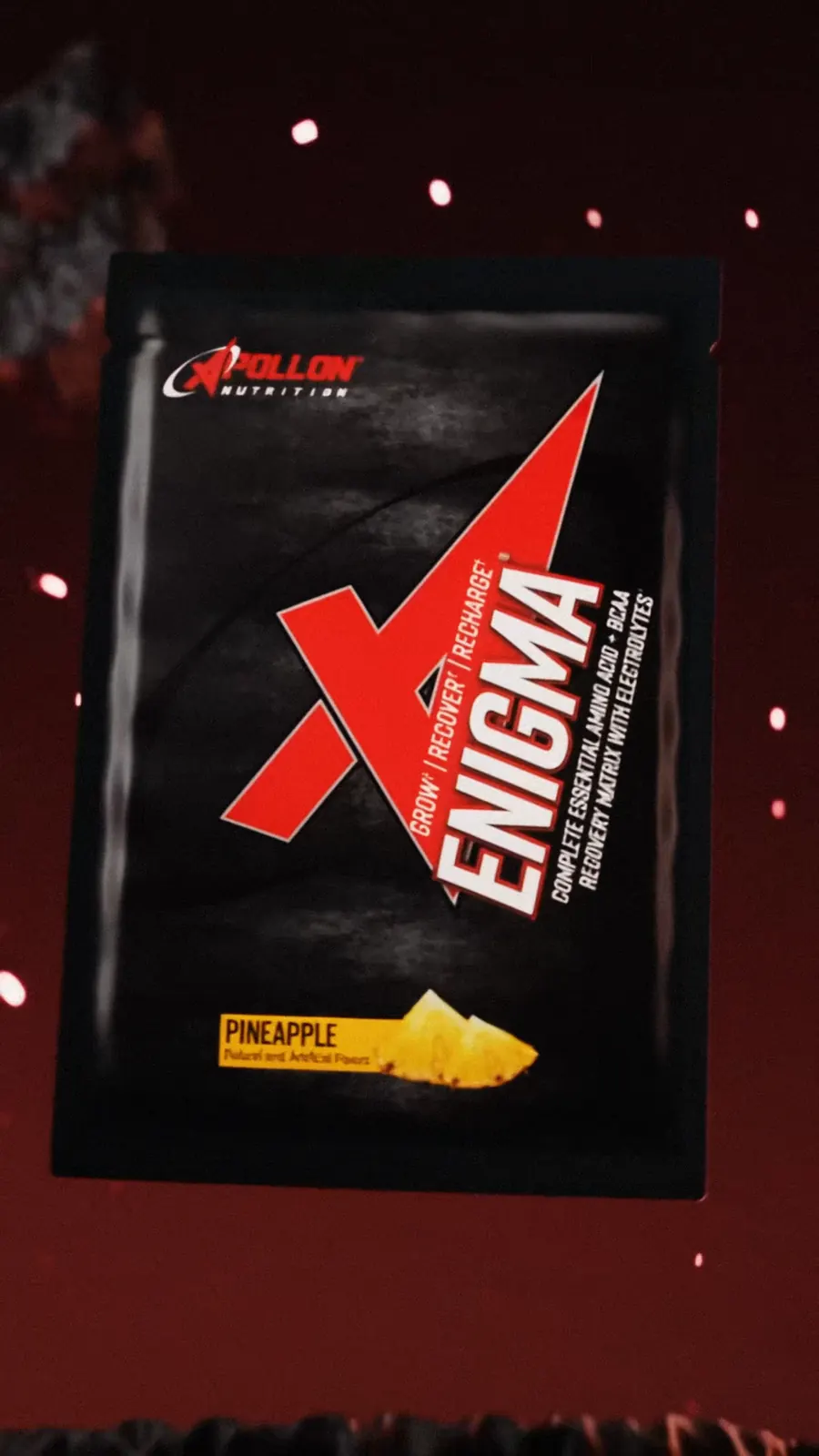 A black Enigma amino sachet in pineapple hovers above dark rock with red sparks.