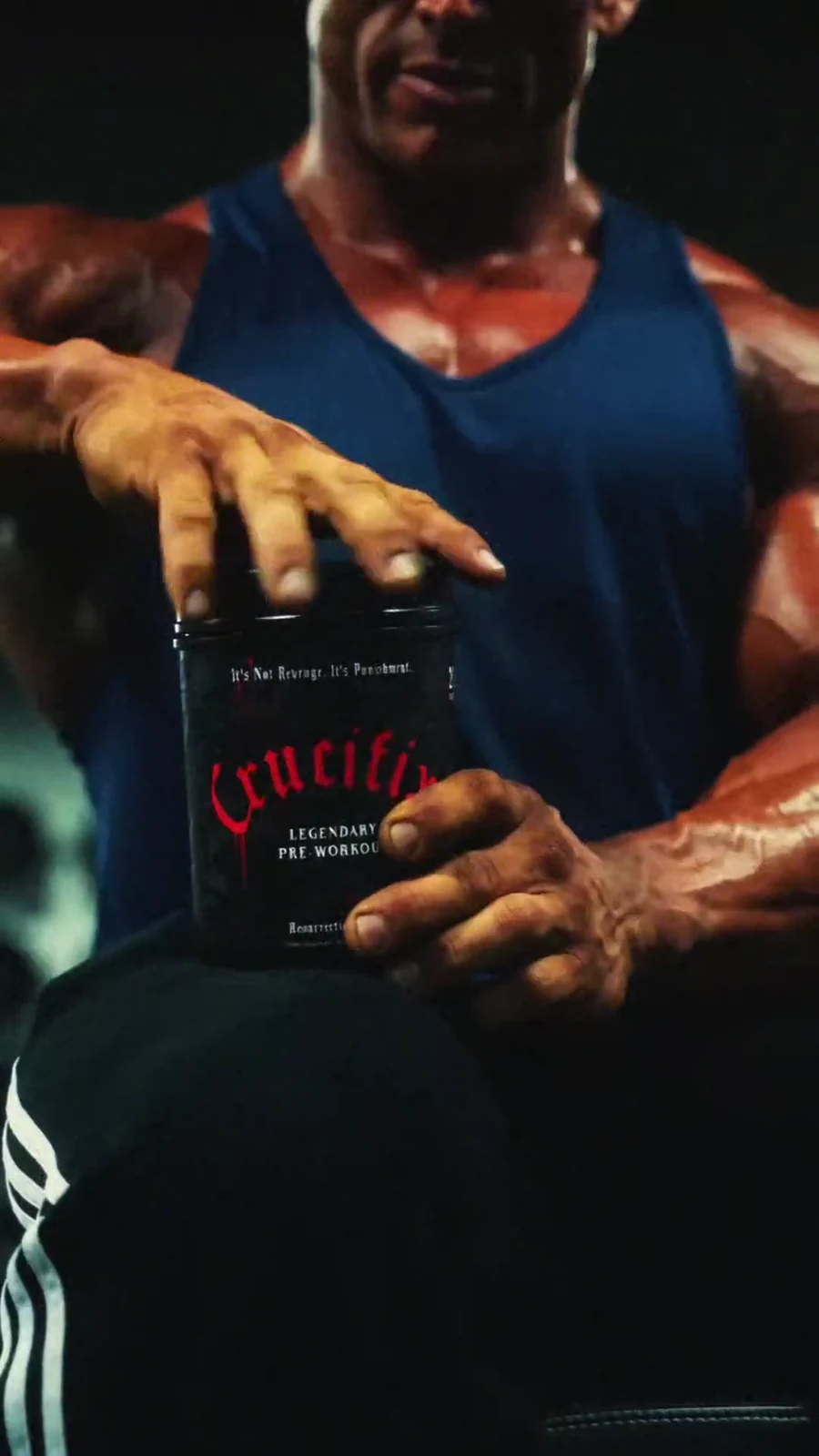 Lee Priest grips a black Crucifix pre-workout tub with its red label facing camera.