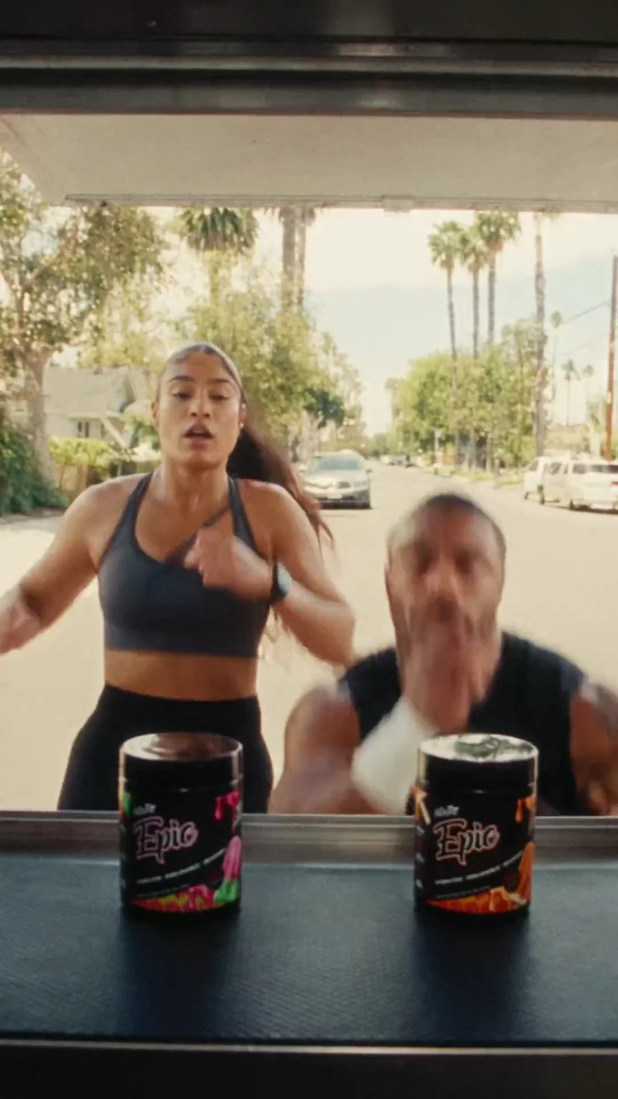 Two athletes arrive at a serving window behind watermelon and mango Epic hydration tubs.