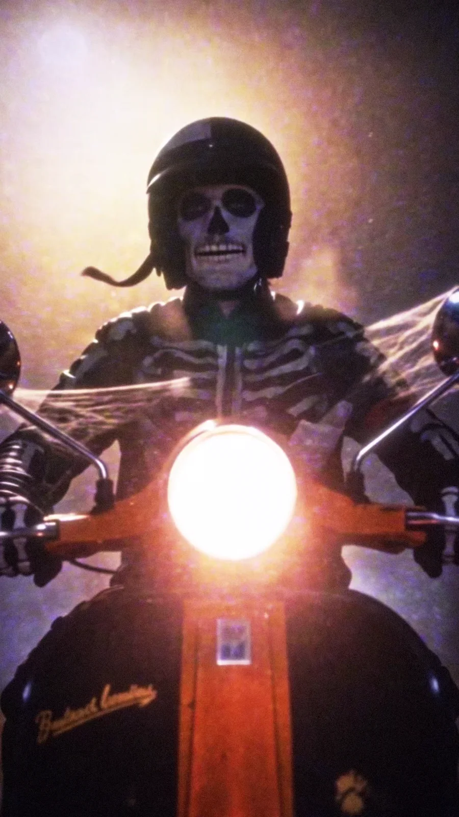 Courier in a skeleton costume and open-face helmet rides a cobwebbed orange scooter through fog, headlight blazing.