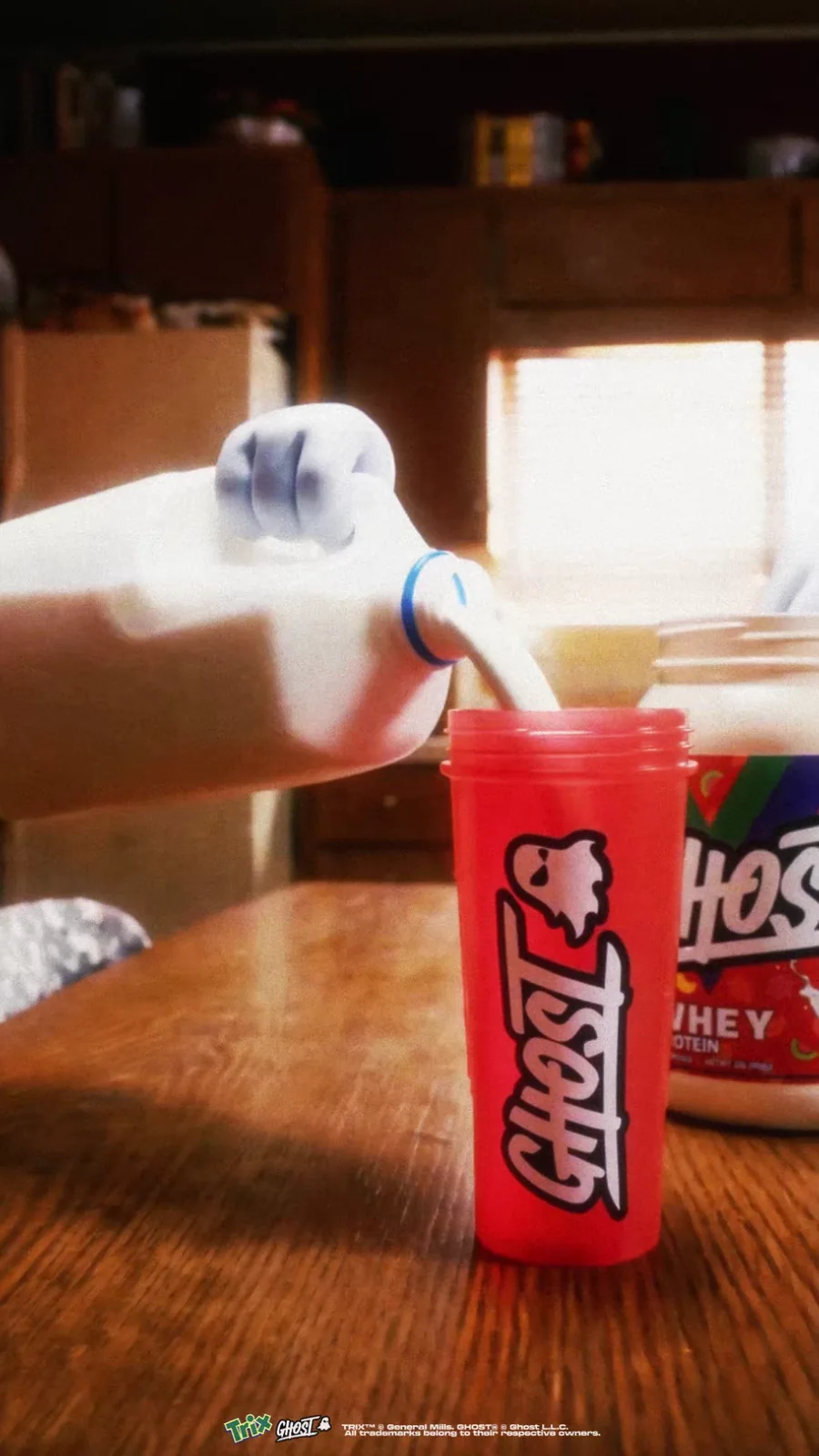 Milk pours into a red Ghost shaker beside a Ghost x Trix whey tub in a warm kitchen.