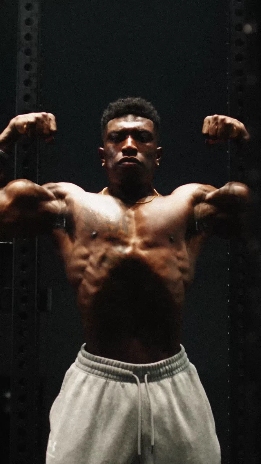 A shirtless bodybuilder holds a front double-biceps pose beneath hard overhead light in a dark gym.