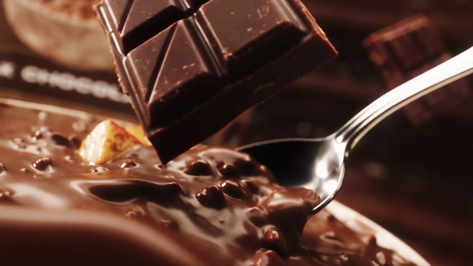 Dark chocolate bar hovering over glossy chocolate creamy rice beside a silver spoon.