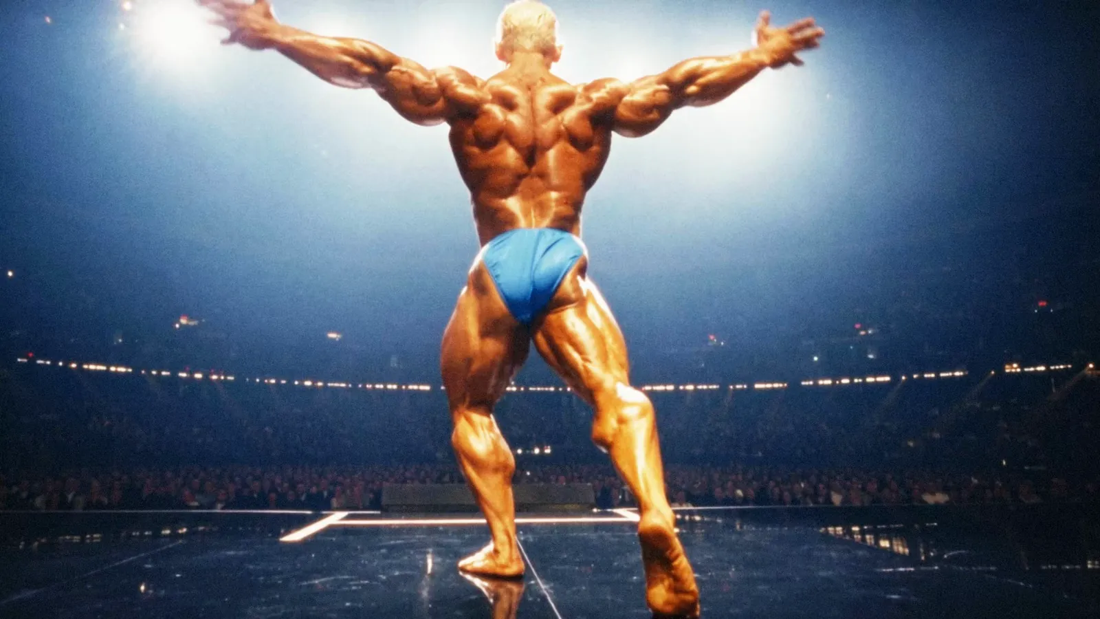 A bodybuilder hits a back double biceps under blue arena lights on a competition stage.