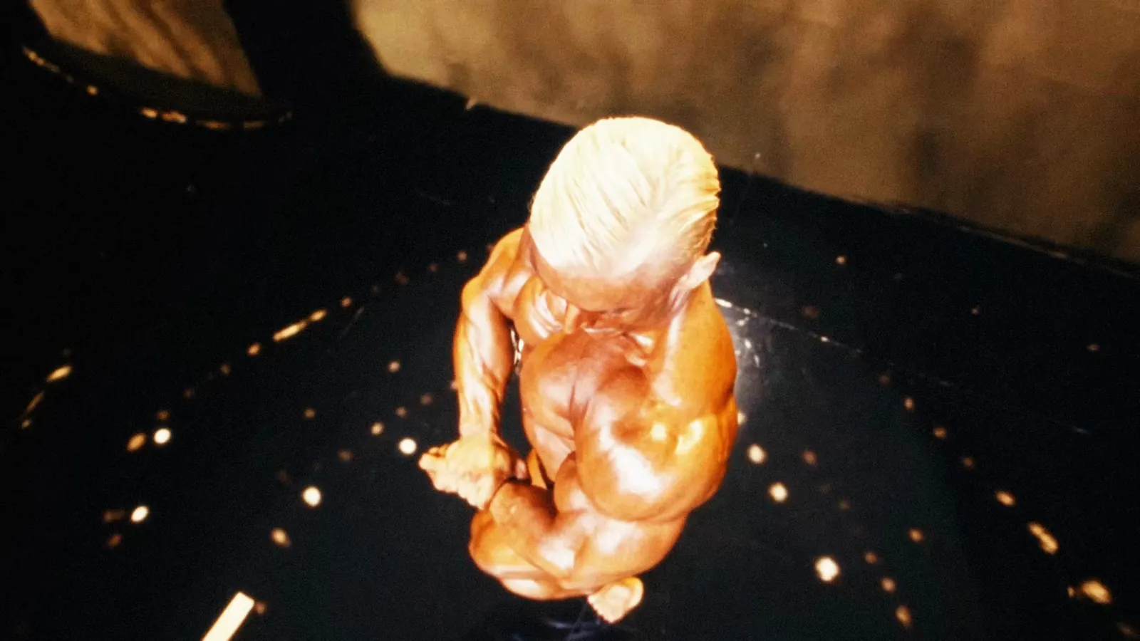 Top-down view of a bronzed bodybuilder hitting a side chest pose on a dark stage.