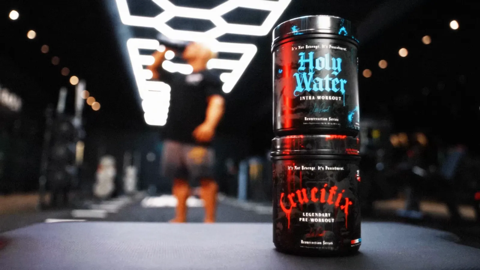 Holy Water and Crucifix tubs stacked in the foreground while Lee Priest drinks from a shaker in the gym.