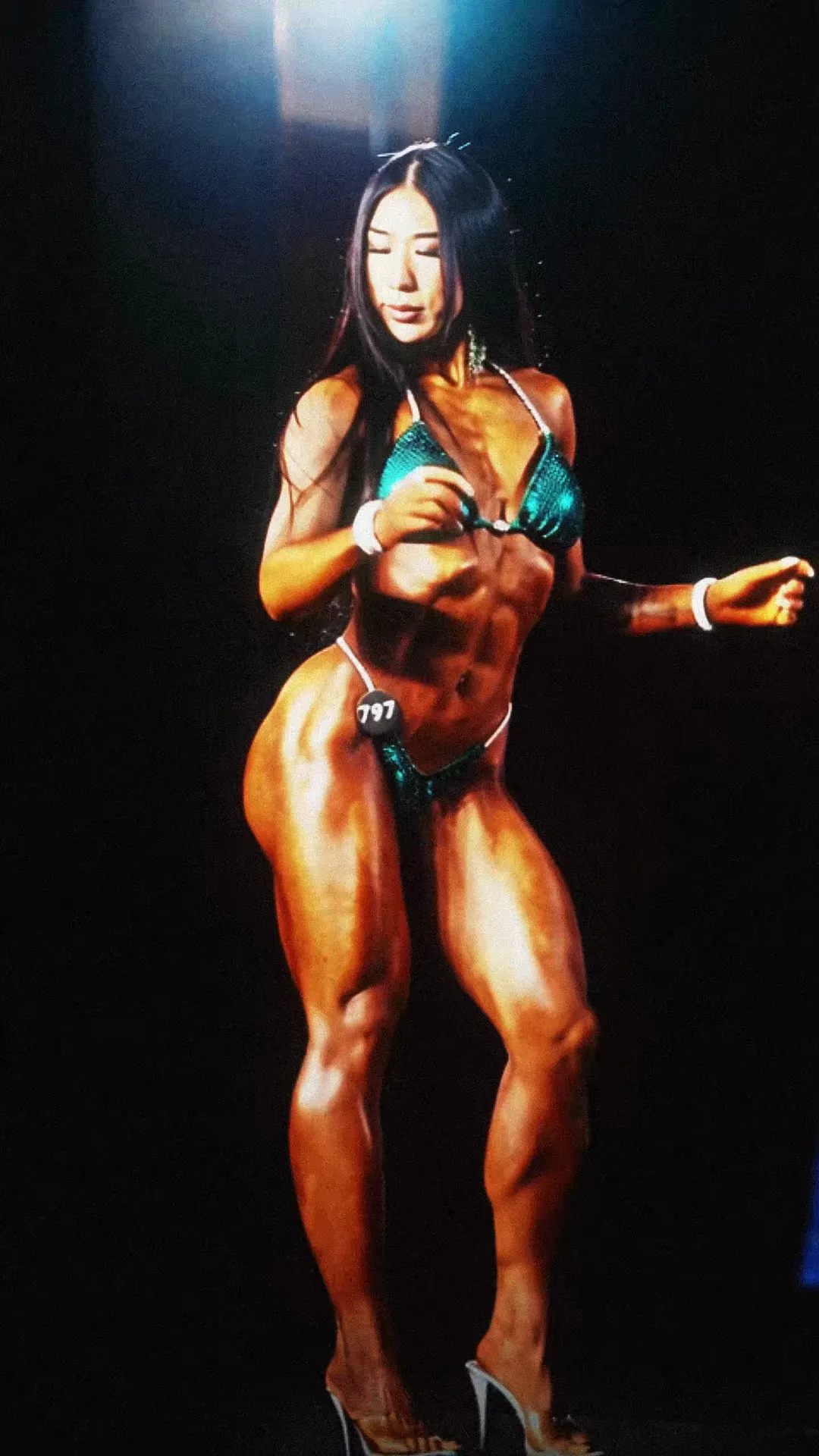 A bikini competitor poses under blue stage lights.