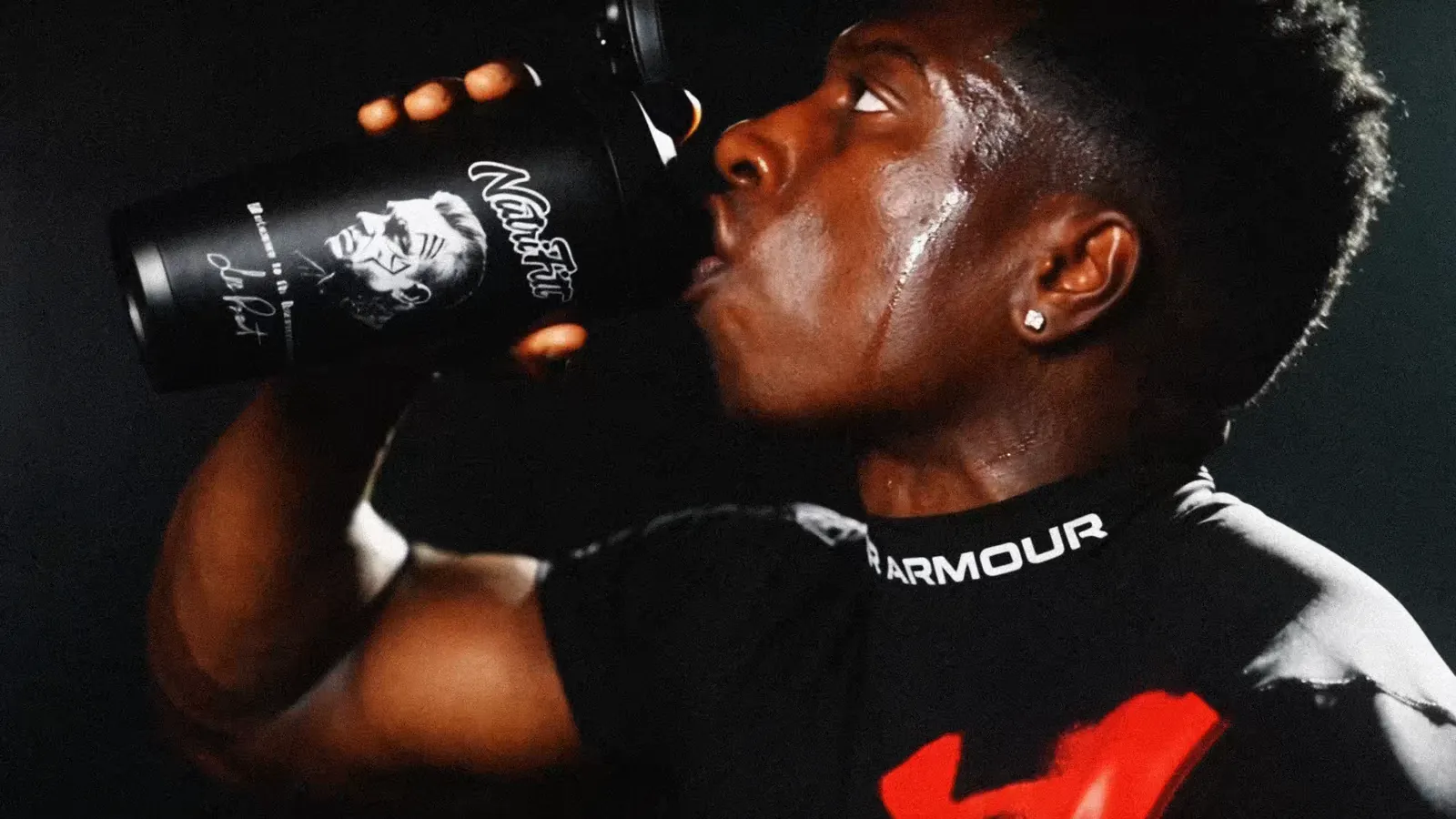 TheOneCay drinks from a black NutriFitt shaker in a dark gym, sweat on his face.