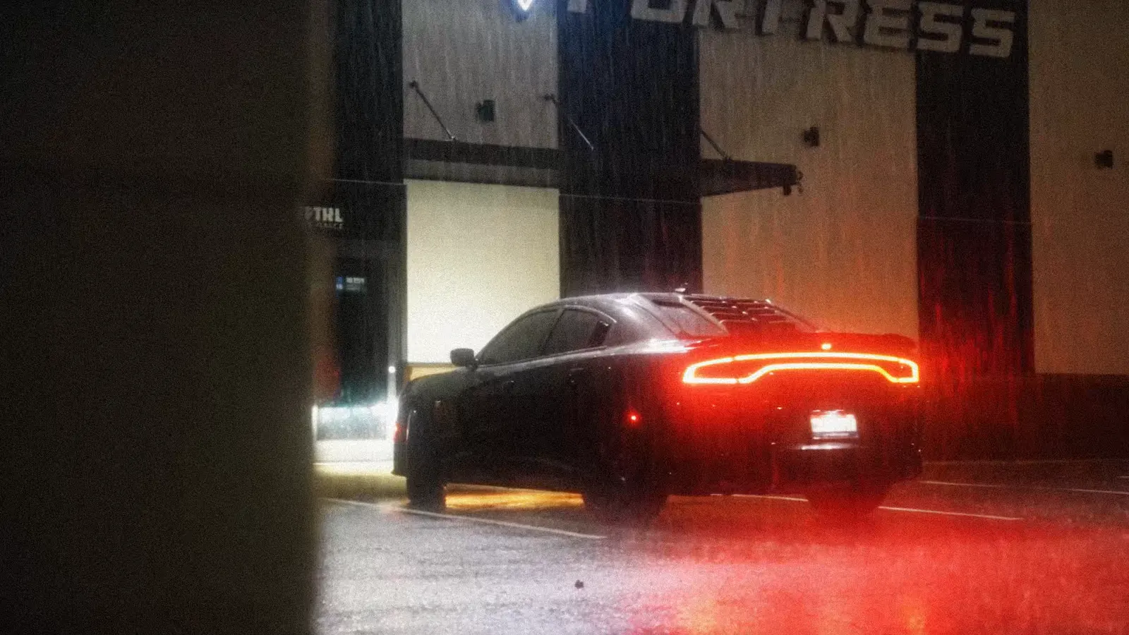 A black muscle car sits in the rain outside the Fortress gym, taillights glowing red.
