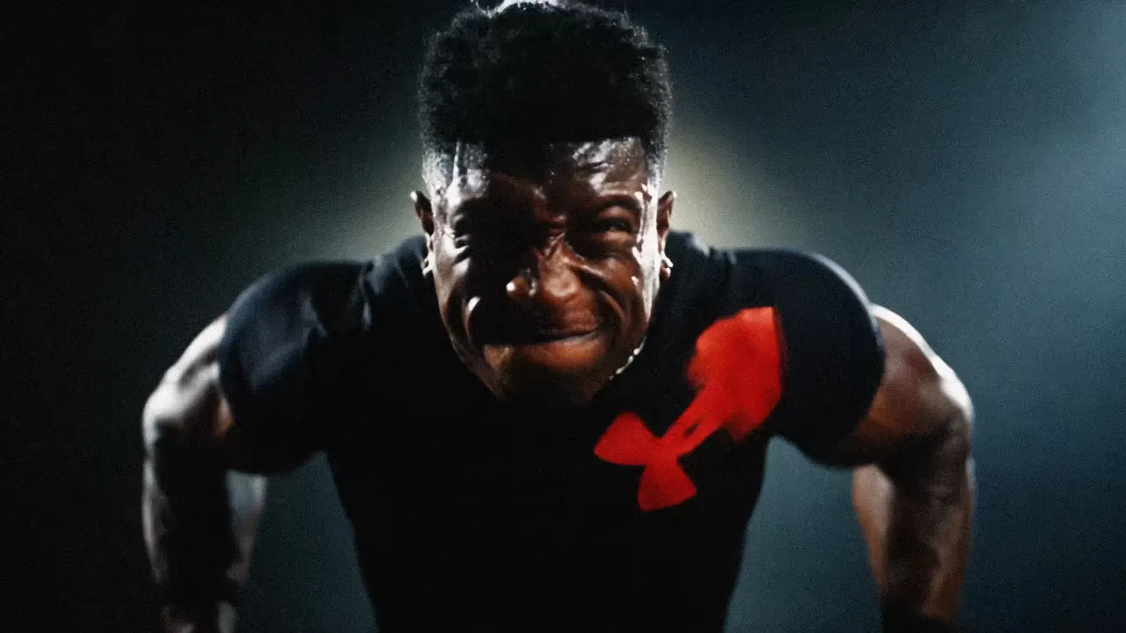 TheOneCay braces on the gym floor in a black Under Armour shirt, lit by a single hard light.