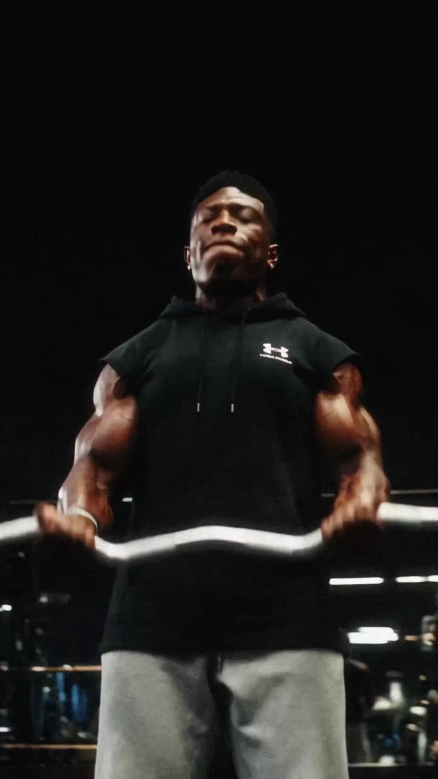 TheOneCay curls a loaded bar beneath hard top light during the strength sequence.