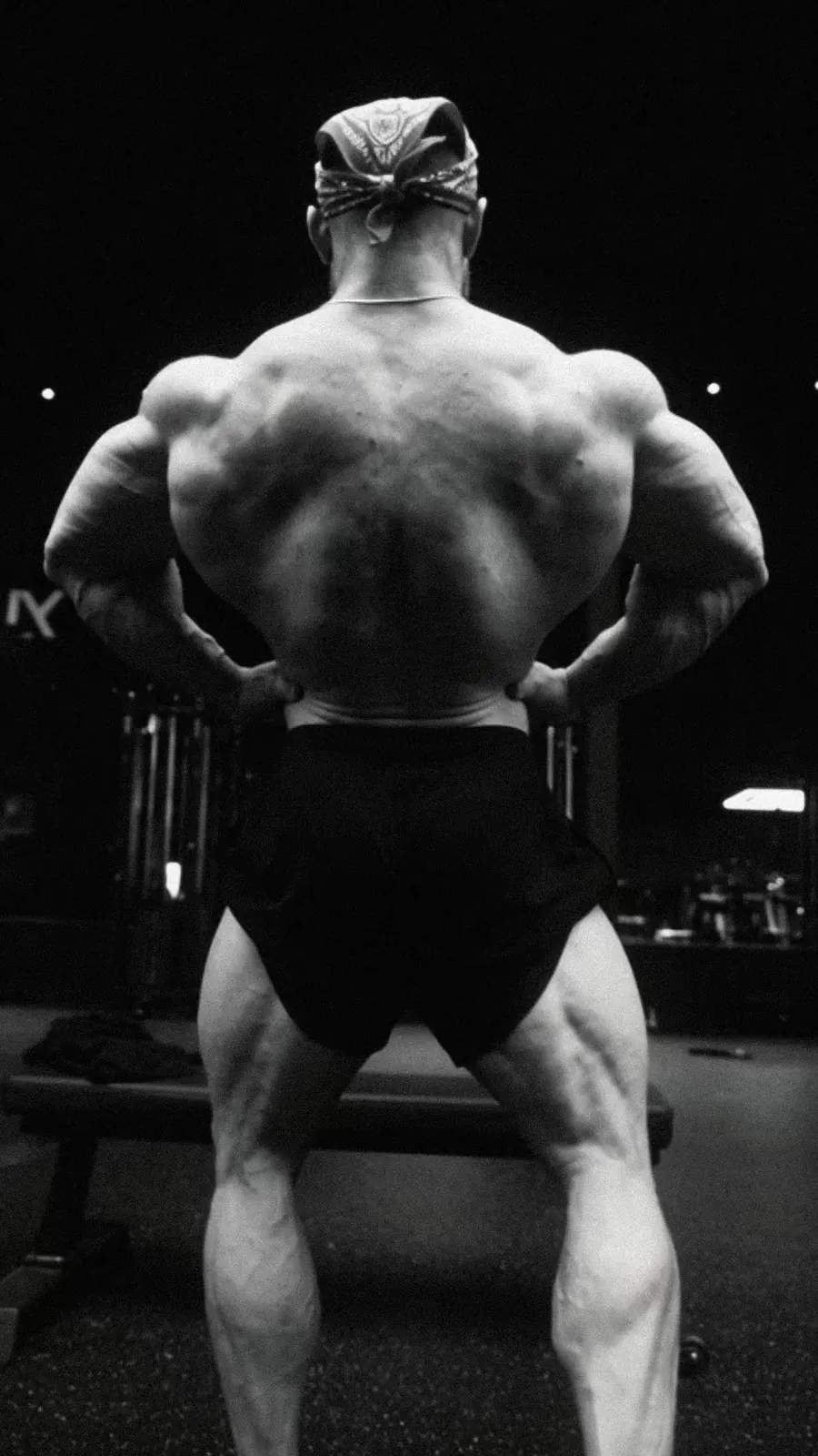 A bodybuilder hits a rear double biceps in black and white under gym lights.