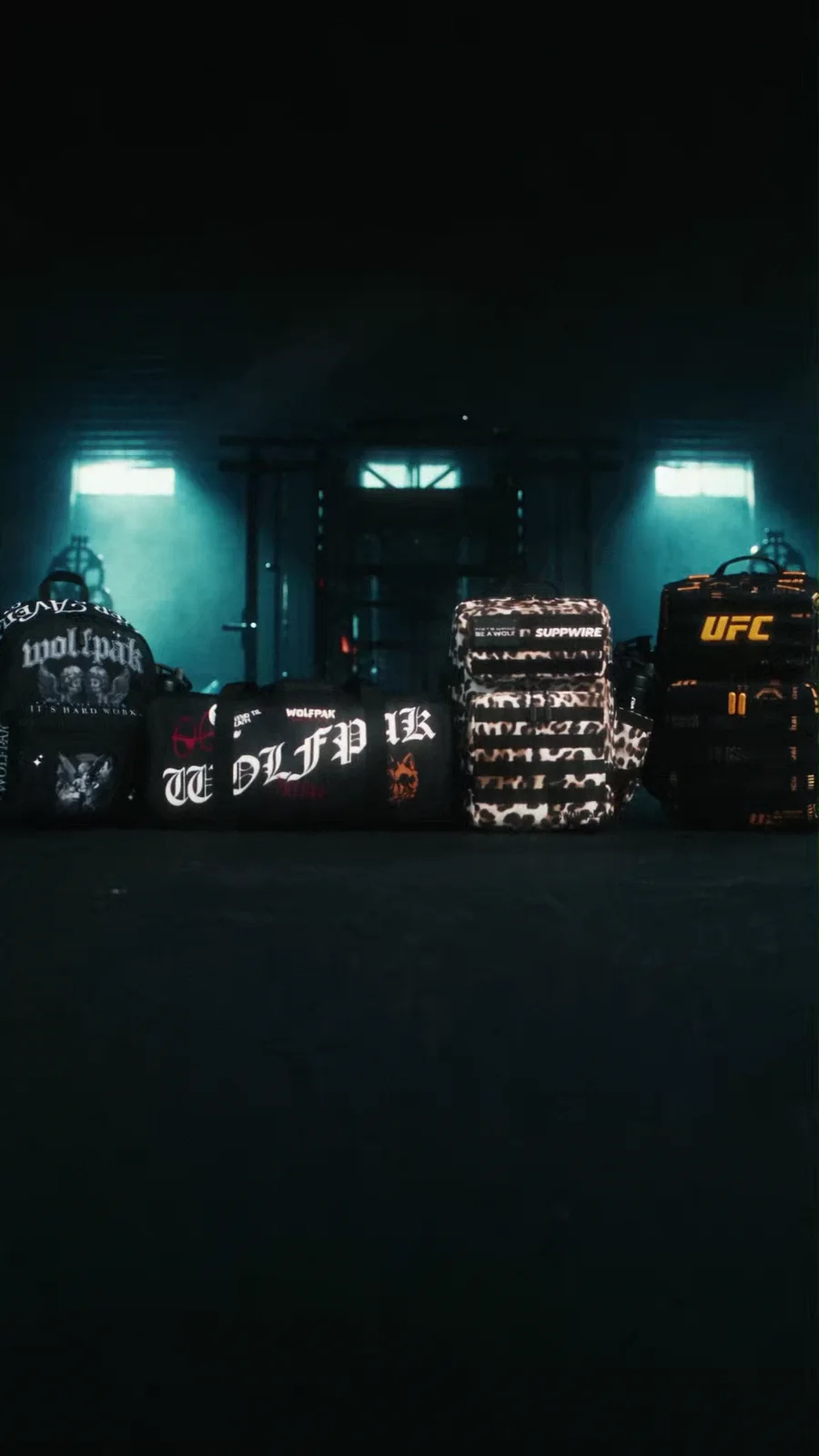 A lineup of WOLFpak bags, including a leopard bag with a Suppwire patch and a black UFC bag, staged beneath teal gym lights.
