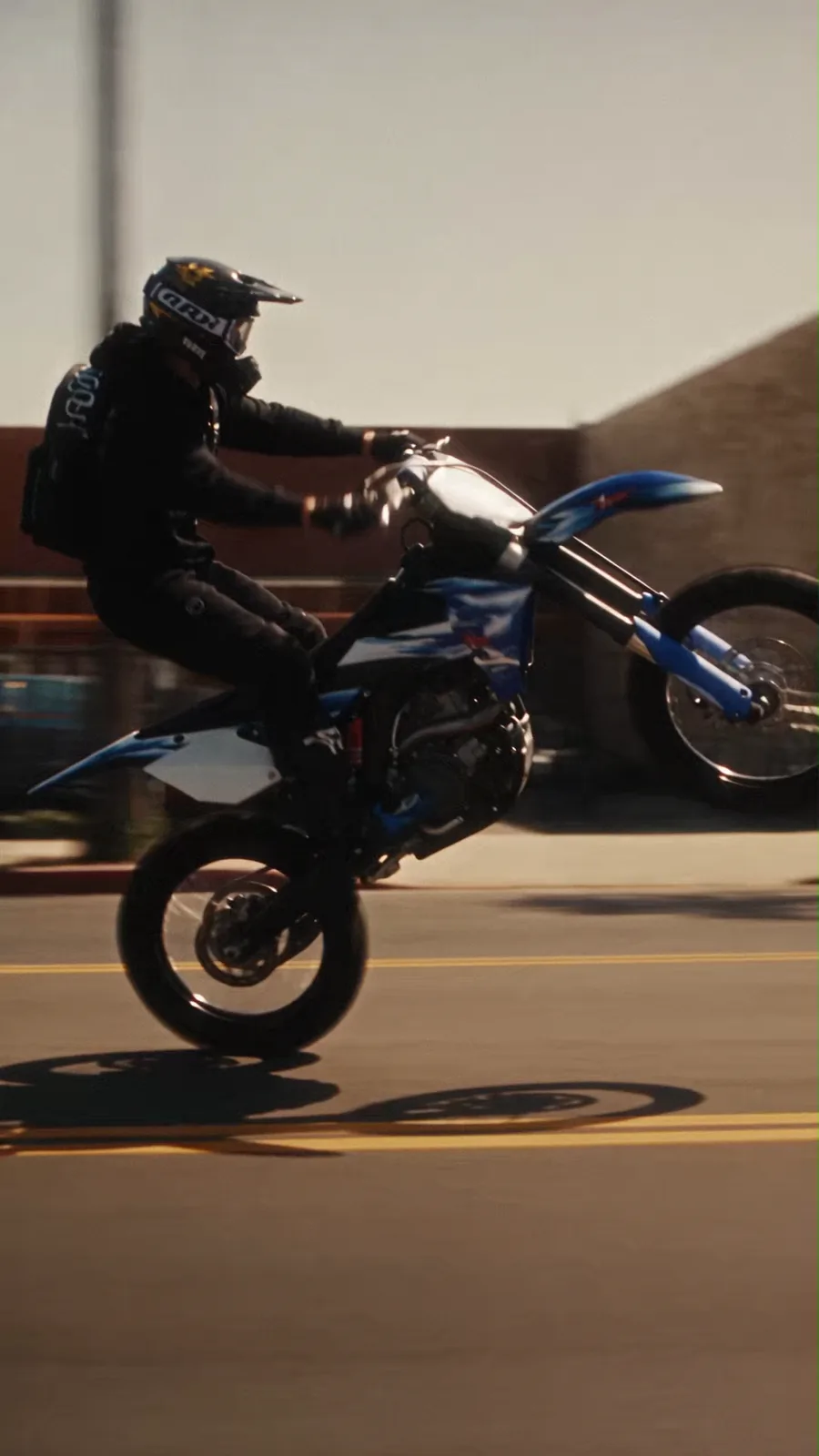 Side tracking view of a rider performing a wheelie on a blue motocross bike.