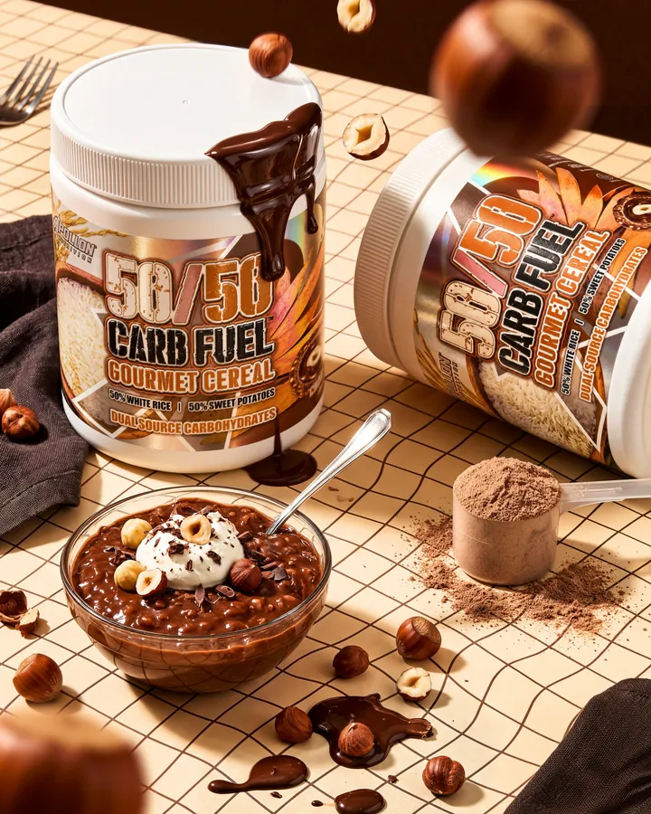Two 50/50 Carb Fuel tubs on a checkered kitchen counter, one on its side, beside a chocolate hazelnut cereal bowl, hazelnuts and a scoop of powder.