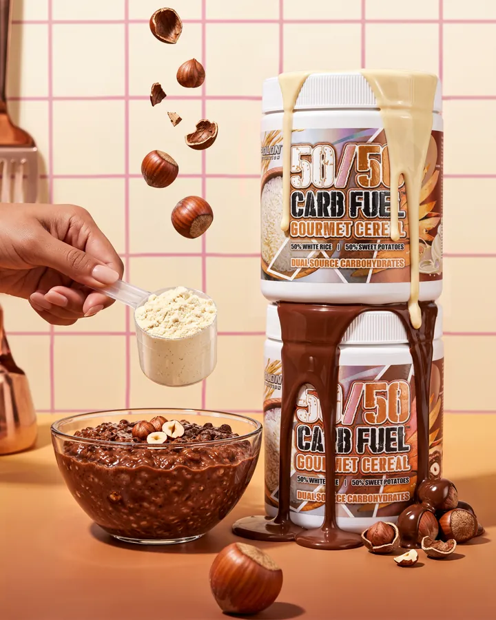 A hand holds a scoop of powder beside two stacked 50/50 Carb Fuel tubs with chocolate running between them.