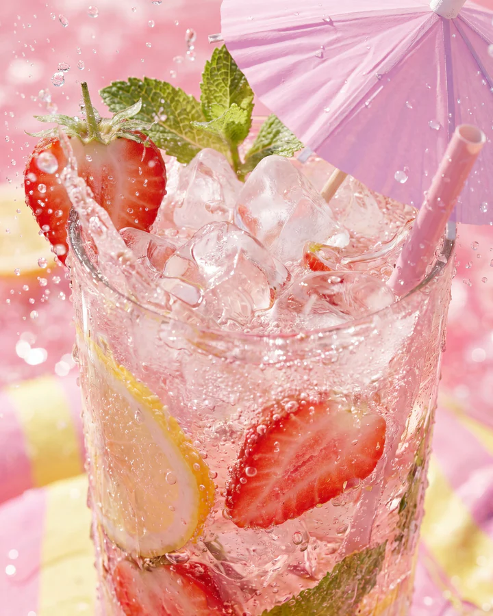 Pink strawberry lemonade with ice, lemon, mint and a paper umbrella fills a sunlit close-up.