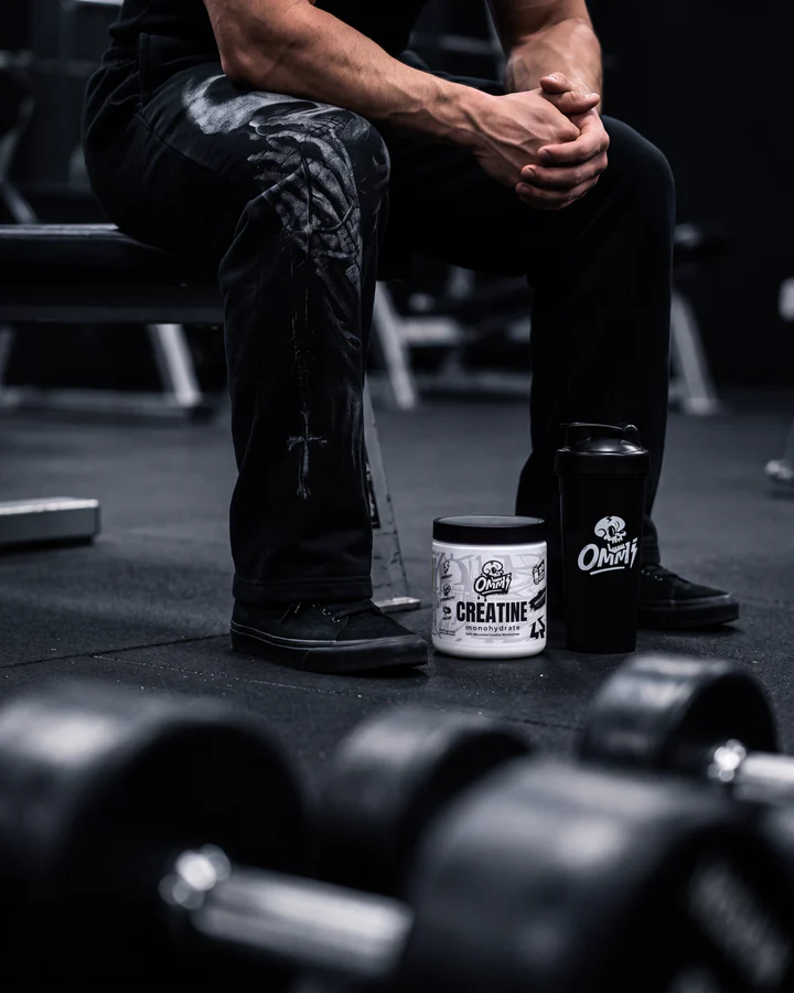 Seated athlete rests in a dark gym with an Ommi Creatine tub positioned near his feet.