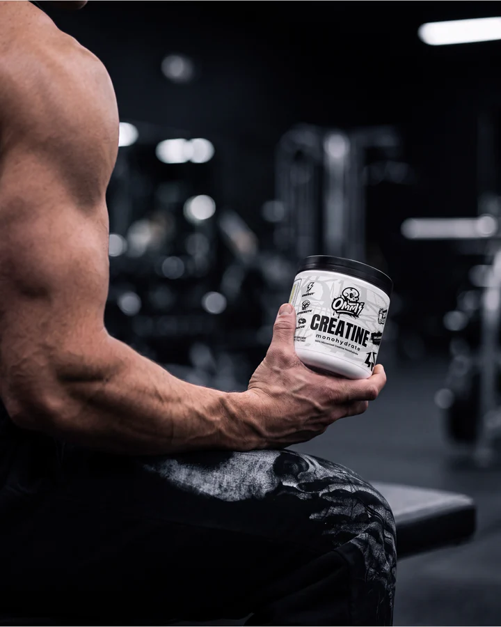 Muscular athlete holds an Ommi Creatine tub toward camera on a gym bench.