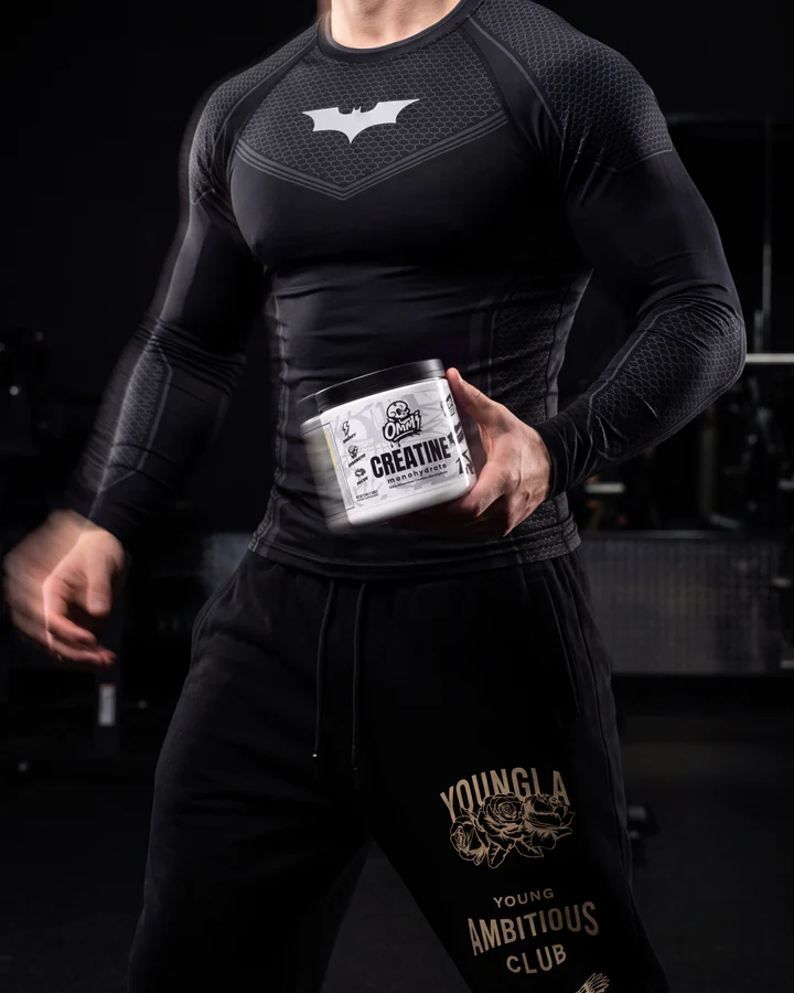 Female strength athlete stands in a dark gym holding an Ommi Creatine tub at her waist.