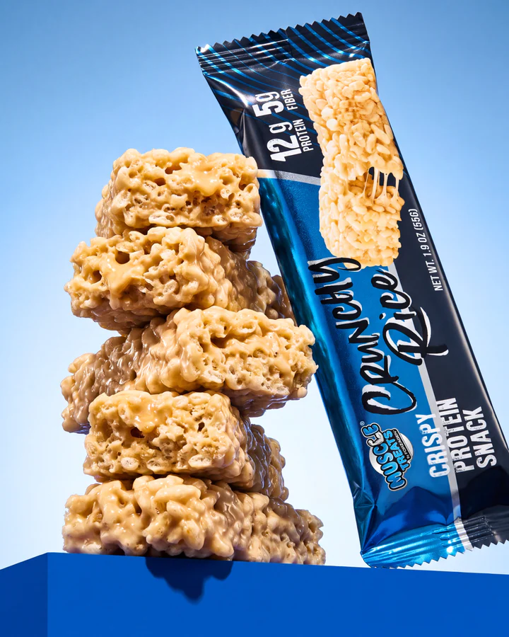 Muscle Treats Crunchy Rice wrapper stands beside a tower of glossy rice-crisp protein snack pieces on a sky-blue set.