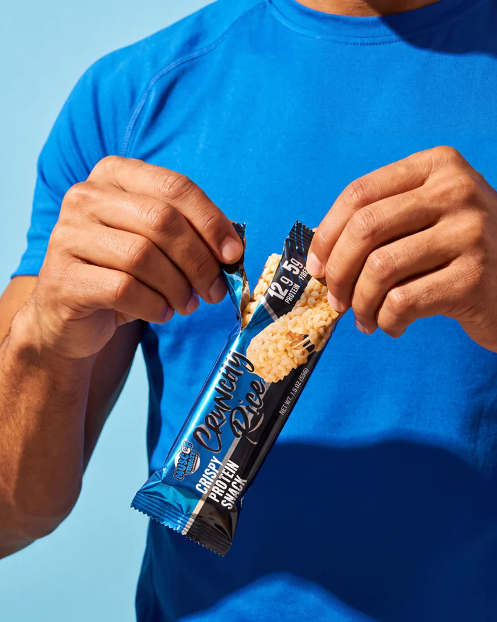 A person in a bright-blue shirt opens a Muscle Treats Crunchy Rice wrapper with both hands.
