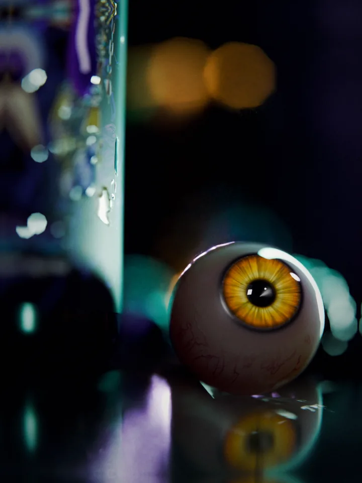 Macro of an eyeball with a yellow iris on wet stone with its reflection, the wet side of the FLAME tub out of focus on the left and warm lights behind.