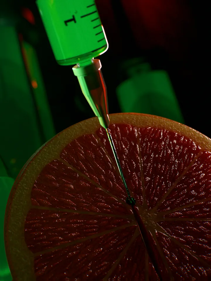 Macro of a syringe needle pushed into the cut face of a red grapefruit, the green dose in the barrel above.