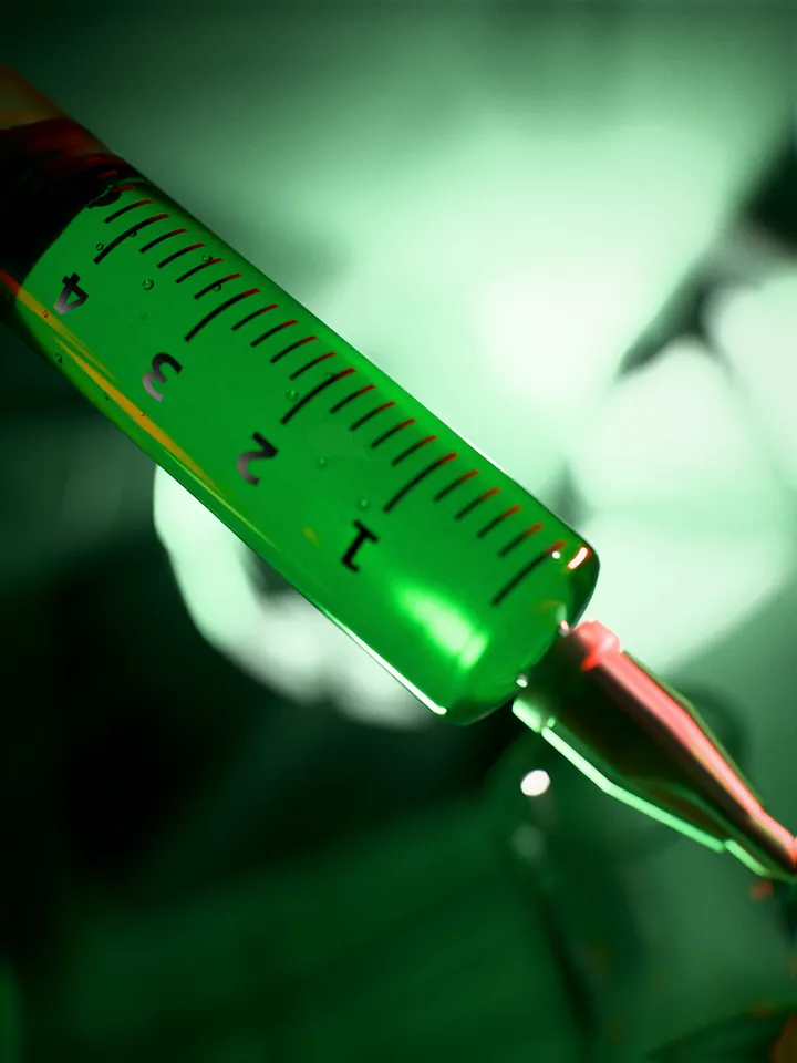 Macro of a syringe barrel with a printed scale and green liquid inside, the lit fan blurred behind it.
