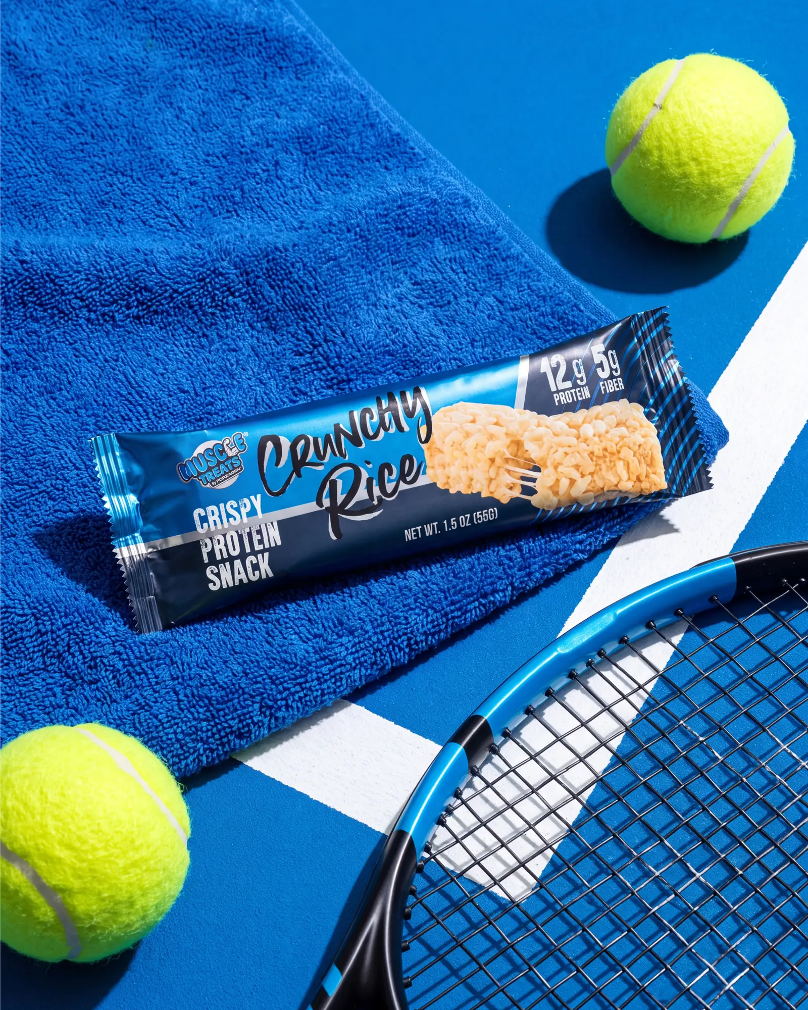 Crunchy Rice protein snack staged with a blue tennis court, racket, towel and tennis balls.