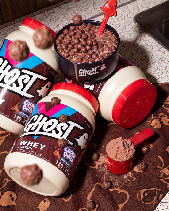 Overhead product composition with three Ghost Cocoa Puffs whey tubs, a cereal bowl, milk and a red protein scoop.