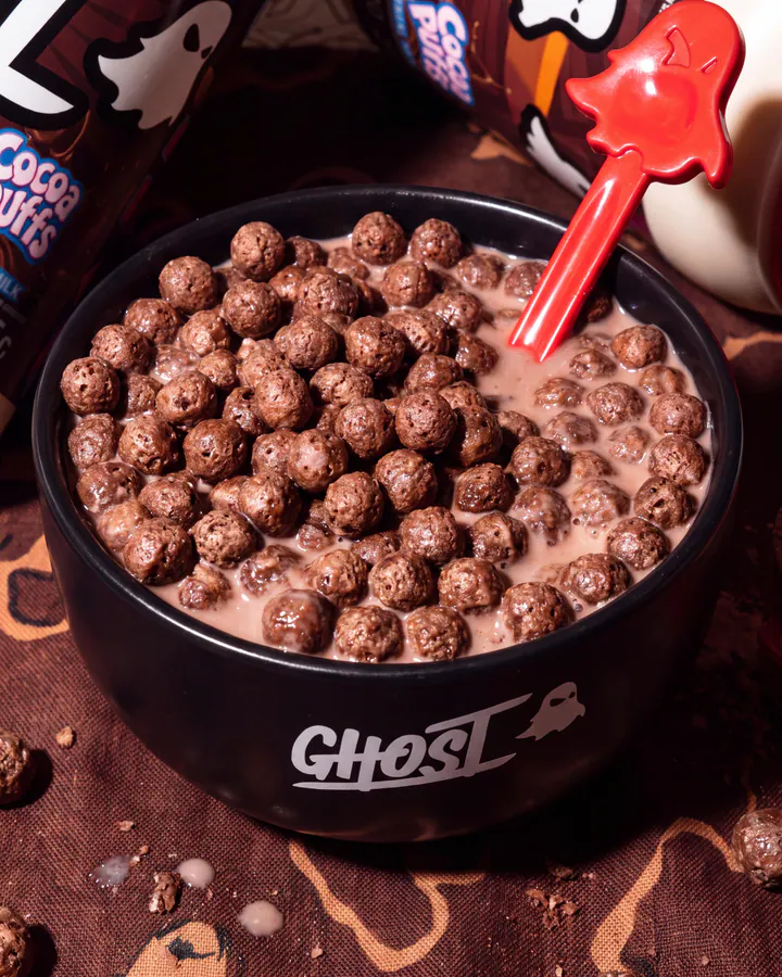 Chocolate cereal and milk in a black Ghost bowl beside Ghost Cocoa Puffs whey packaging.