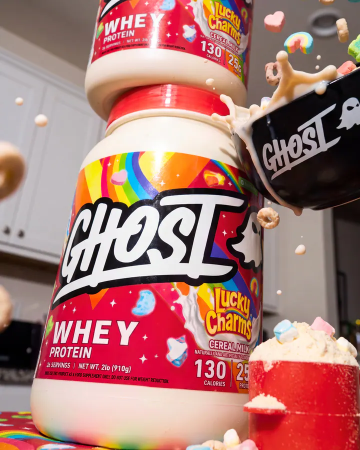 Two Ghost Lucky Charms whey tubs surrounded by cereal pieces, a black bowl and a red protein scoop.