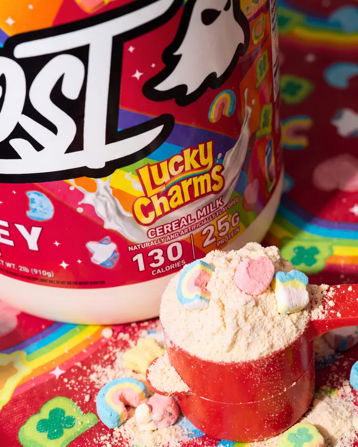 Ghost Lucky Charms whey tub and a red scoop of protein powder on a bright cereal-covered table.