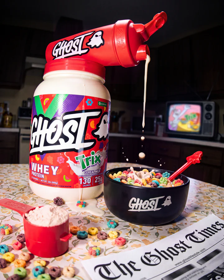 Ghost x Trix whey tub stands with a red Ghost scoop, black cereal bowl, colorful cereal and newspaper texture.