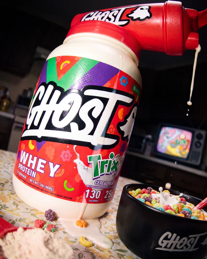 Angled Ghost x Trix whey tub sits beneath a red scoop with a thin milk drip beside a cereal bowl.
